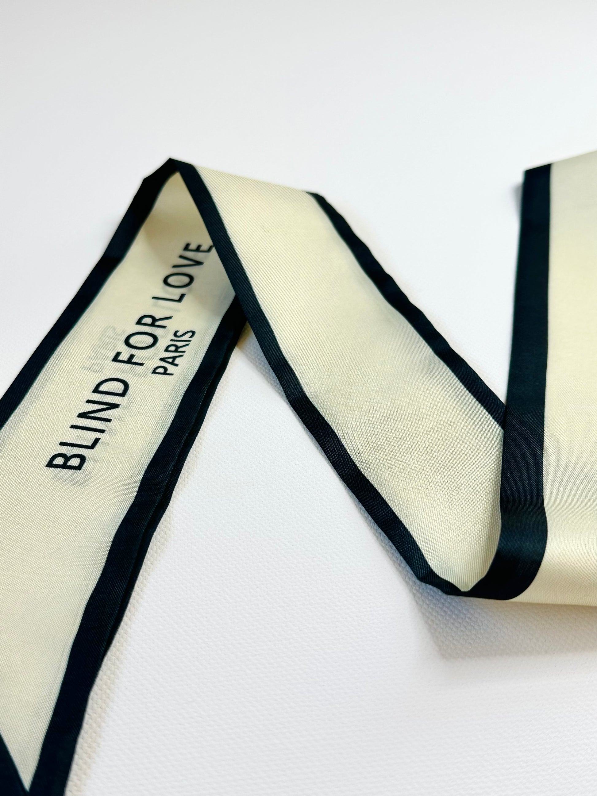 Cream-colored ribbon scarf with black trim and 'Blind for Love Paris' text on a white background
